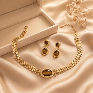 Könnte beinhalten: Eine goldfarbene Halskette und ein passendes Paar Ohrhänger, jeweils mit einem ovalen Tigerauge-Edelstein. Die Halskette besteht aus einer mehrsträngigen geflochtenen Kette mit einem zentralen ovalen Stein, während die Ohrringe einen einzelnen Stein tragen.