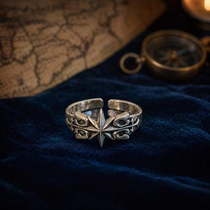 Könnte beinhalten: Ein silberfarbener Metallring mit offenem Banddesign und einem zentralen achtstrahligen Sternmotiv. Die Oberfläche des Rings zeigt komplizierte, erhabene Schnörkelmuster, die vom Stern ausgehen und mit einer dunklen, oxidierten Textur versehen sind.