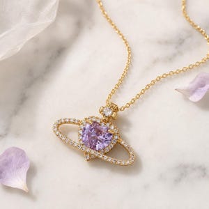 Könnte beinhalten: Eine goldfarbene Halskette mit einem Anhänger in Form eines Planeten mit Ring. In der Mitte des Anhängers befindet sich ein herzförmiger, hellvioletter Edelstein, der von kleinen klaren Steinen umgeben ist. Ein kleiner Stein sitzt oben.