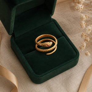 Könnte beinhalten: Ein goldfarbener Metallring in Form einer gewundenen Schlange mit einer texturierten, schuppenartigen Oberfläche. Der Schlangenkopf ist mit zwei kleinen, eingelassenen Edelsteinen in Grün und Rot versehen, die als Augen auf dem Ringband sitzen.