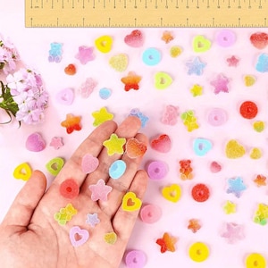 May include: A collection of small, colourful, sugar-coated gummy sweets in shapes of stars, hearts, rings, and bears, scattered across a light pink surface next to a wooden ruler measuring up to 22 centimetres in length.