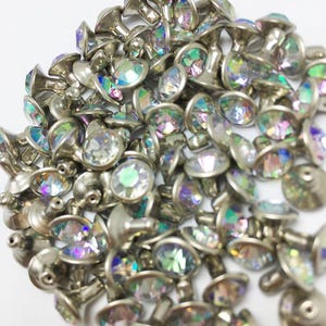 May include: A collection of silver-toned metal rivets featuring circular, faceted iridescent crystals set in the center. Each rivet has a smooth metallic base and a pronged back, displaying a reflective, multi-colored shimmer across the surface.