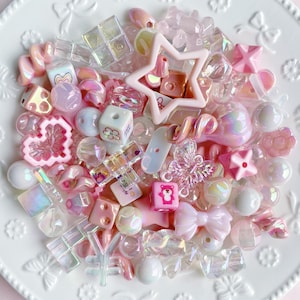 Op de afbeelding: Een verzameling diverse knutselkralen in tinten roze, wit en iriserend transparant. De mix bevat vormen zoals sterren, harten, strikjes, blokjes met beren- en bloemenprints, vlinders en gedraaide spiralen, allemaal met een glanzende afwerking.
