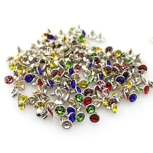 May include: A collection of silver-toned metal rivets featuring circular, faceted rhinestones in a variety of vibrant colors including red, blue, green, yellow, and pink. Each rivet has a small metal post on the back for attachment.