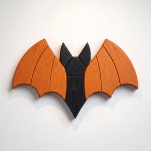 May include: A wooden wall decoration shaped like a bat with a black central body and two large, orange-stained wings. The piece features visible wood grain patterns and segmented wing sections, creating a stylized representation of a bat.
