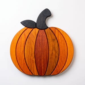 May include: A pumpkin-shaped wooden board constructed from multiple vertical segments of wood in varying shades of orange, red, and brown. The board features a dark black wooden stem and a single black leaf attached at the top center.