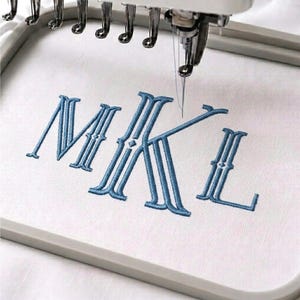 Op de afbeelding: Een borduurmachinenaald boven een witte stoffen ring, die de blauwe monogramletters M, K en L borduurt. De letters hebben een decoratief schreeflettertype met een dubbele omtreklijn en kleine ruitvormige accenten op elke verticale lijn.
