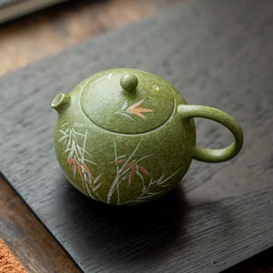May include: A round, textured green ceramic teapot featuring a hand-painted bamboo design in white and copper tones. The teapot has a matching lid with a small knob, a curved handle, and a short spout, showcasing a speckled finish throughout.