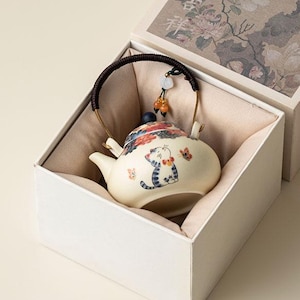 May include: A cream-colored ceramic teapot with a hand-painted blue and grey tabby cat and butterfly motif, featuring a dark brown woven handle, a blue lid knob, and a decorative tassel, presented in a square box with floral artwork.