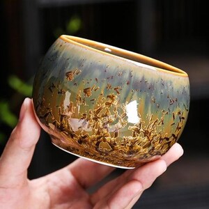 Puede incluir: Una pequeña taza de té de cerámica redonda con un esmalte reactivo que presenta un degradado de tonos verde apagado, marrón y azul oscuro. La superficie exterior muestra un patrón texturizado y cristalino que recuerda a hojas dispersas o restos orgánicos.