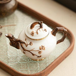 Könnte beinhalten: Eine weiße Keramik-Teekanne mit einem braunen, astförmigen Griff, Ausguss und Deckelknauf, verziert mit gemalten braunen Blumenmotiven. Die Teekanne steht auf einem rechteckigen Holztablett mit einem strukturierten Glasboden neben einem kleinen braunen Gefäß.