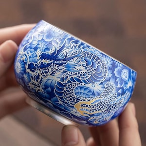 Könnte beinhalten: Eine kleine Keramikschale mit einer detaillierten blau-weißen Drachenillustration vor einem dichten Blumenhintergrund. Das Drachendesign enthält feine weiße Linien und dezente goldene Akzente auf den Schuppen, die die gesamte Außenfläche bedecken.