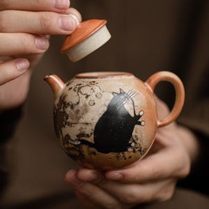 Op de afbeelding: Een ronde keramische theepot met een gespikkeld oranje en bruin glazuur, voorzien van een zwarte kat-illustratie op de zijkant. De theepot heeft een kleine tuit, een gebogen handvat en een bijpassend afneembaar deksel met een lichte onderkant.