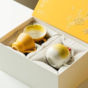 May include: A gift box containing two ceramic mugs. One mug is solid yellow with a textured finish, and the other is a round, off-white mug with a yellow and green gradient. The box lid is bright yellow with white floral line art and Chinese text.