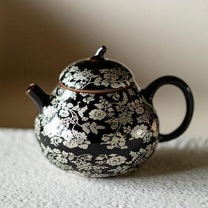 Puede incluir: Una tetera redonda de cerámica de color marrón oscuro con un patrón floral blanco detallado y repetitivo en toda su superficie. La tetera incluye una tapa a juego con un pequeño asa puntiaguda, un pico curvo y un asa lateral arqueada.