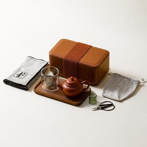 May include: A tea set featuring a rectangular brown storage box with a wide elastic band, a wooden tray, a small brown teapot, a clear glass cup, a grey cloth with a bamboo graphic, a small grey drawstring pouch, a green glass, and metal scissors.
