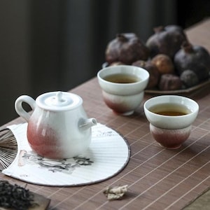 May include: A white ceramic teapot and two matching tea cups featuring a gradient red-to-white glaze. The teapot has a rounded body, a curved handle, and a lid. The tea cups are small and bowl-shaped. A decorative fan sits beneath the teapot.