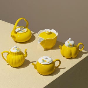 Puede incluir: Una colección de cinco teteras de cerámica de color amarillo brillante y un cuenco con tapa. Cada pieza tiene un acabado brillante, algunas con tapas blancas decoradas con pequeños motivos florales amarillos y hojas verdes, con formas variadas.