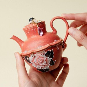 Può includere: Una teiera color corallo con una rosa bianca dipinta a mano con foglie nere sul corpo. Il coperchio è decorato con una piccola perla nera, un ciondolo a forma di fiore bianco e un cristallo rosa a goccia pendente. Una catenella dorata con minuscoli cristalli rosa avvolge la parte centrale, accentuata da dettagli a linee dipinte in bianco.