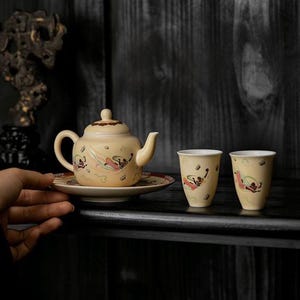 Puede incluir: Una tetera de cerámica de color amarillo claro con tapa a juego y dos tazas pequeñas, decoradas con figuras pintadas de colores con túnicas fluidas y motivos de nubes. La tetera descansa sobre un platillo circular y tiene un acabado esmaltado.