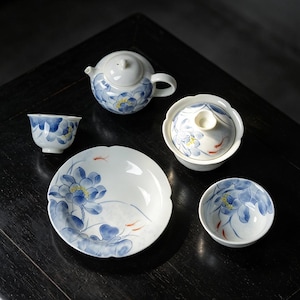 May include: A white ceramic tea set featuring a hand-painted blue lotus flower pattern and small orange fish accents. The set includes a teapot, a lidded bowl, two small tea cups, and a matching saucer, all decorated with consistent floral motifs.