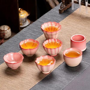 May include: A set of six small ceramic tea cups with a gradient pink and white finish. Four cups feature a scalloped, petal-like rim, while two are cylindrical. The cups are filled with amber-coloured tea, showcasing a smooth, glazed texture.