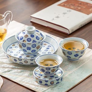 May include: A ceramic tea set featuring a white base with a repeating pattern of blue and gold floral motifs. The set includes a lidded gaiwan on a matching saucer, two small tea cups, and one saucer, all filled with a light amber tea liquid.