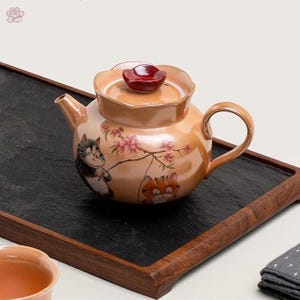 Puede incluir: Una tetera de cerámica color melocotón con tapa festoneada y pomo rojo oscuro. El cuerpo presenta ilustraciones de dos gatos y una rama florida. La tetera tiene un asa curva y un pico corto, con un acabado brillante en toda la superficie.