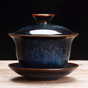 Puede incluir: Un juego de té gaiwan de cerámica compuesto por un cuenco con tapa y un platillo a juego. El conjunto presenta un esmalte moteado en azul profundo y negro con un acabado brillante y detalles en marrón en los bordes de la tapa, el cuenco y el platillo.
