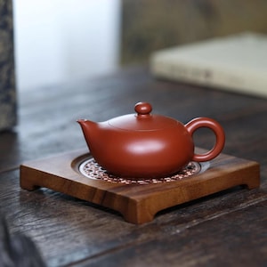 Peut inclure: Une théière en argile brun rougeâtre lisse avec un corps arrondi, un bec verseur incurvé et un petit bouton sur le couvercle. La théière repose sur une base carrée en bois dotée d'un insert métallique circulaire couleur cuivre perforé.