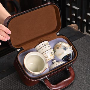 以下が含まれることがあります： 茶色のレザー調キャリングケースには、ジッパーとハンドルが付いており、陶器のティーセットが収納されています。セットには、ティーピッチャー、3つの小さなカップ、蓋付きの茶筒、および別の蓋が含まれており、すべてに風景画が描かれています。