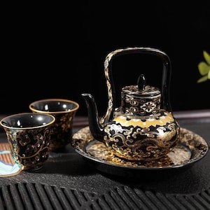 Puede incluir: Un juego de té de cerámica negra que incluye una tetera con asa alta arqueada, tapa y dos tazas a juego. El conjunto está decorado con intrincados patrones de remolinos dorados y blancos y bordes geométricos sobre una superficie negra brillante.