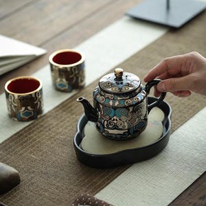 May include: A ceramic teapot and two matching cups featuring a black, white, and turquoise blue swirling pattern. The teapot has a gold-toned knob on the lid, and the cups have a vibrant red interior. The set rests on a black, cloud-shaped tray.