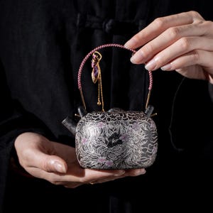 Puede incluir: Una tetera de cerámica negra con intrincados grabados plateados de un zorro rodeado de motivos en espiral y pequeñas flores de cerezo rosas. La tetera tiene un pico curvo y un asa trenzada rosa con un dije dorado colgante.