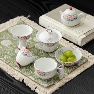 Puede incluir: Un juego de té de cerámica blanca con delicados motivos florales en tonos coral, azul y dorado. El conjunto incluye una tetera pequeña, un gaiwan con tapa, una copa con pie, un cuenco con uvas verdes y una taza con asa de cola de pez.