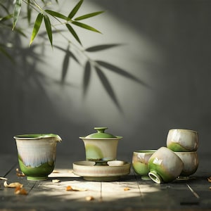May include: A ceramic tea set featuring a pitcher, a lidded gaiwan on a saucer, and four small cups. The pieces have a gradient glaze transitioning from a deep forest green at the base to a creamy off-white and light brown at the top rim.