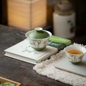 May include: A ceramic tea bowl with a matching lid and a separate small tea cup, both featuring a white base with green accents and gold-painted bamboo leaf motifs. The lidded bowl sits atop a book, while the smaller cup rests on a textured mat.