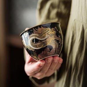 May include: A black ceramic tea cup featuring a detailed gold dragon illustration that wraps around the exterior. The dragon design includes intricate scales and flowing lines, with stylised wave patterns at the base of the cup and a gold rim.