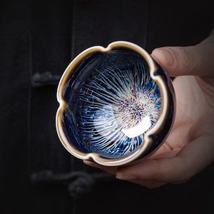 May include: A small ceramic tea bowl with a scalloped rim and a deep blue glaze featuring a radiating white and brown crystalline pattern that resembles a starburst or flower, with a smooth, glossy finish on the interior and exterior surface.