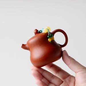 May include: A small, reddish-brown clay teapot with a rounded body, a short spout, and a curved handle. The lid features a small knob and is decorated with a dangling ornament made of colourful green, yellow, and blue beads on a thin cord.