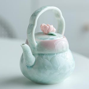 Op de afbeelding: Een keramische theepot met een licht mintgroen glazuur en een kristallijn patroon. De theepot heeft een gebogen handvat, een korte tuit en een deksel bekroond met een gebeeldhouwde lichtroze bloemknop. Het bovenlichaam heeft een zachte roze tint.