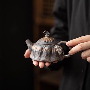 May include: A dark brown and bronze-toned ceramic teapot with a textured, ribbed surface resembling a gourd. The teapot features a curved spout, a lid with a small stem-like handle, and a side handle adorned with a small, detailed leaf accent.