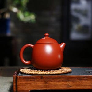 May include: A round, terracotta-colored teapot with a smooth, matte finish, featuring a curved handle, a short spout, and a lid with a small knob handle. The teapot sits on a circular, woven rattan coaster.