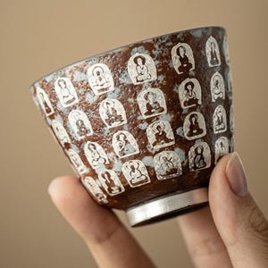 Puede incluir: Una taza de té de cerámica con un esmalte texturizado y moteado en marrón y azul claro. La superficie exterior está decorada con un patrón repetitivo de figuras sentadas blancas y estilizadas en marcos arqueados. Base circular metálica.