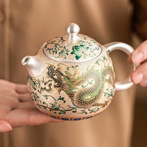 Può includere: Una teiera rotonda in argento con un intricato motivo a drago in smalto nero, verde e oro. La superficie è ricoperta da un fine motivo a filigrana vorticoso, con coperchio abbinato, manico curvo e un piccolo beccuccio. La base presenta accenti blu.