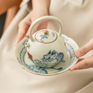 May include: A round ceramic teapot with a high arched handle and a matching saucer, featuring a blue and white dragon illustration, cloud motifs, and a small red square stamp on the side. The teapot has a light cream glaze with a subtle crackle finish.