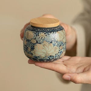 May include: A round ceramic storage jar featuring a dark blue base with a detailed floral pattern in shades of light blue, cream, and gold. The jar is topped with a circular, light-colored wooden lid that has a smooth, natural grain finish.