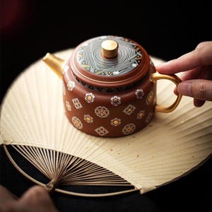 May include: A terracotta-coloured ceramic teapot featuring a gold-toned handle and spout. The body is decorated with a repeating white and gold floral pattern, while the lid displays a black and white geometric swirl design with a gold knob.