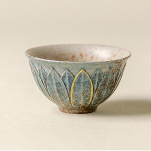 May include: A ceramic tea bowl featuring a textured exterior with carved petal-like shapes in shades of blue and brown. The rim of one petal is highlighted with a gold-coloured accent. The interior surface has a speckled brown and white finish.