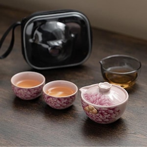 以下が含まれることがあります： ピンクと白の花柄が施された2つの小さな陶器の茶碗と蓋付きの蓋碗からなる茶器セット。琥珀色の茶が入った透明なガラス製のピッチャーが近くにあり、後ろには黒いファスナー付きのキャリングケースが置かれています。